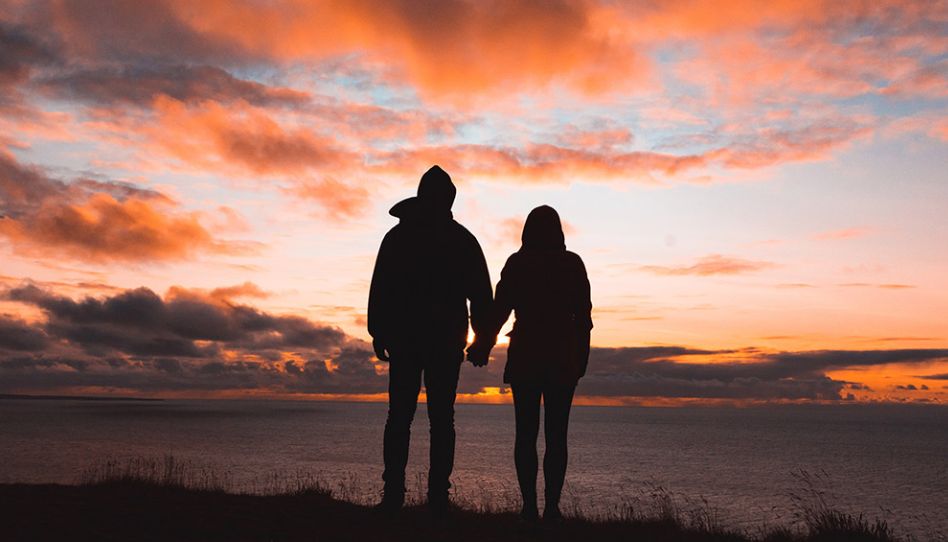 Casal segurando as mãos e contemplando o por do sol.